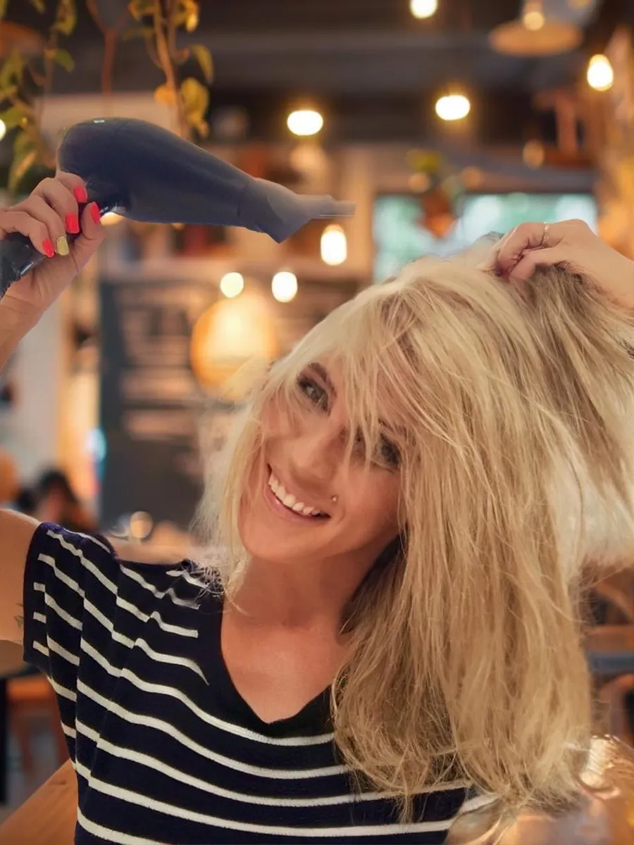 Blonde woman joyfully blow-drying her hair, showing off a carefree, salon-fresh look with playful energy.