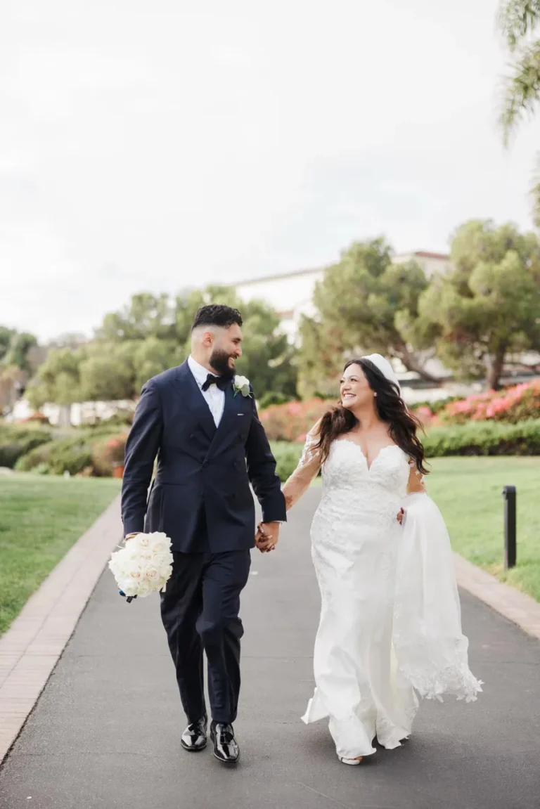 Smiling bride and groom walking hand in hand on their wedding day in a garden setting.