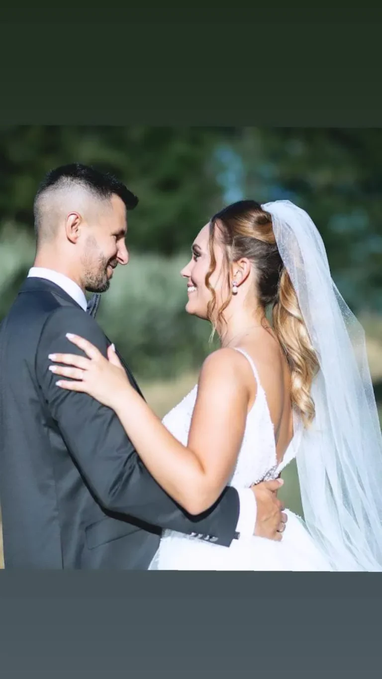 Bride with soft curly updo hairstyle and long veil, smiling at her groom during an outdoor wedding ceremony.