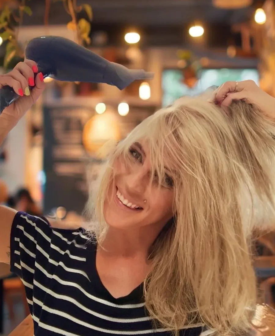 Blonde woman joyfully blow-drying her hair, showing off a carefree, salon-fresh look with playful energy.