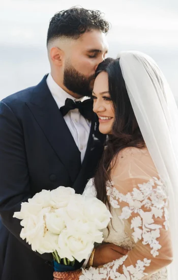 Dettycated bridal moment captured: the bride glows with happiness holding elegant white roses, while the groom gently kisses her forehead a perfect balance of romance, beauty, and elegance.